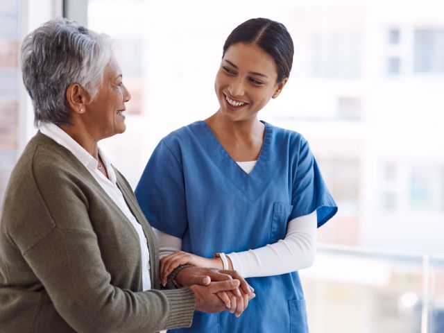 Support, caregiver with senior woman and holding hands for care indoors. Retirement, consulting and professional female nurse with elderly person smiling together for healthcare at nursing home.
