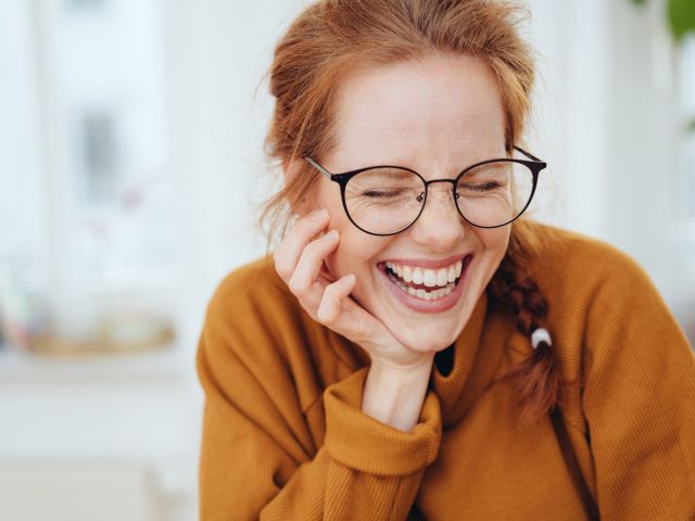 Pretty red-haired girl with pigtail, wearing glasses and orange sweatshirt, laughing with her eyes closed. Close-up front portrait indoors with copy space