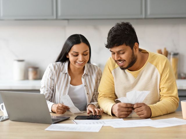 Planning Family Budget. Smiling indian couple hold papers, calculate domestic bills, check household expenses, read documents or receipt from bank, talk about investment and finance, kitchen interior